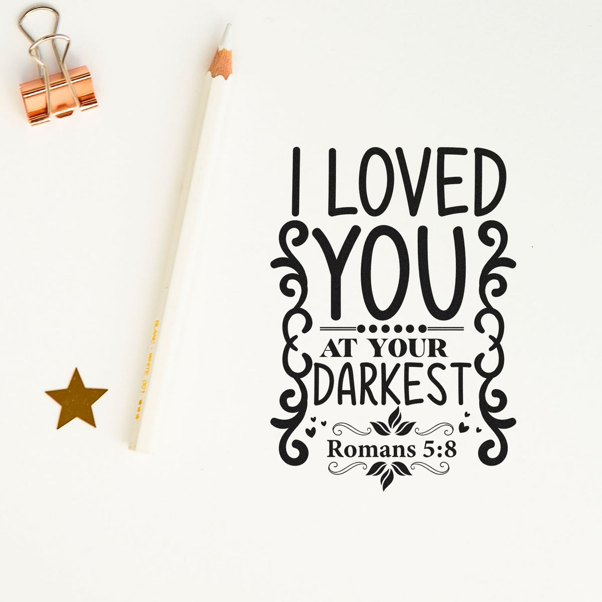 Black decorative stamp reading I loved you at your darkest with Romans 5:8, on white, beside a pencil, gold star and rose gold clip.