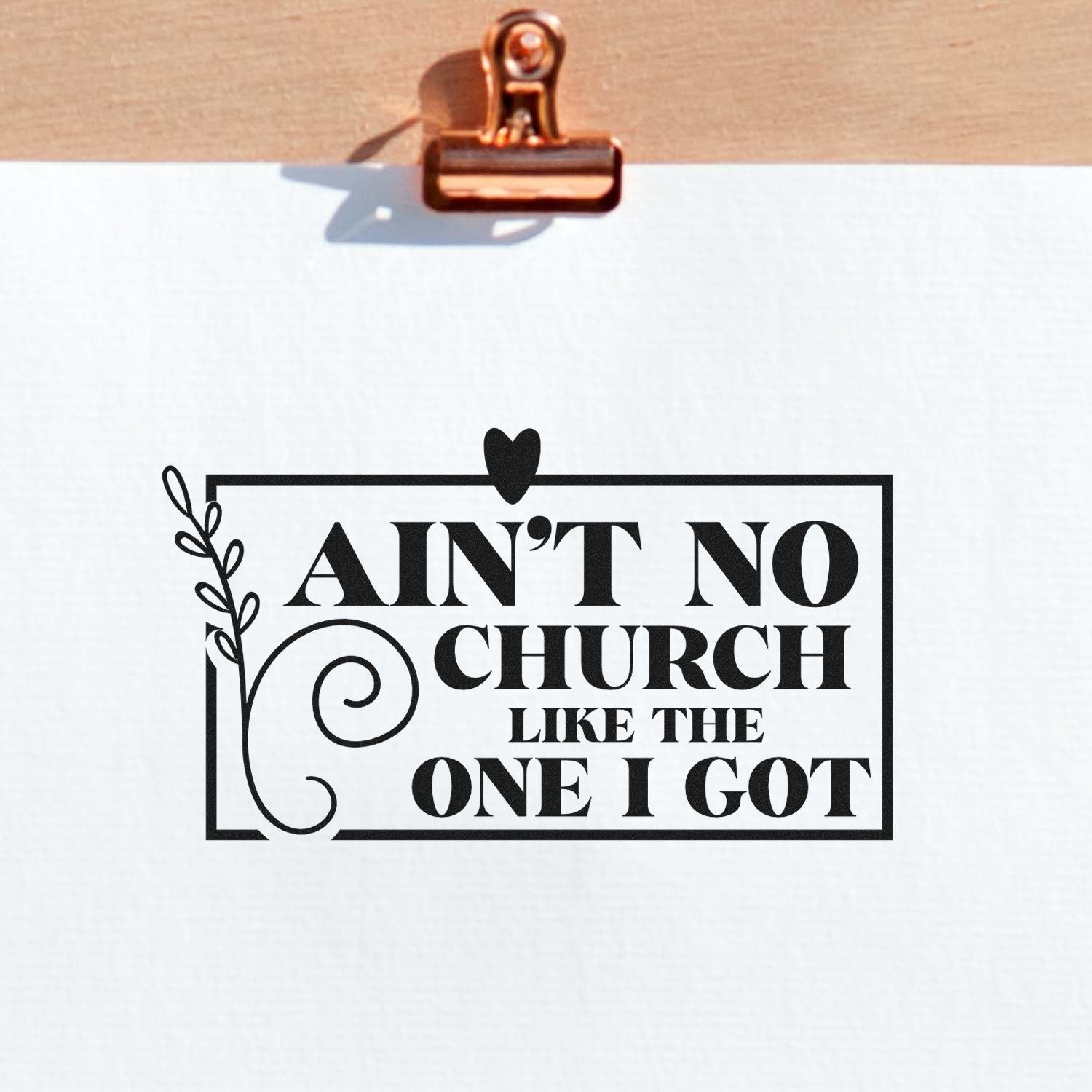 Black decorative stamp seal reading AINT NO CHURCH LIKE THE ONE I GOT on white paper, clipped with a copper clip.
