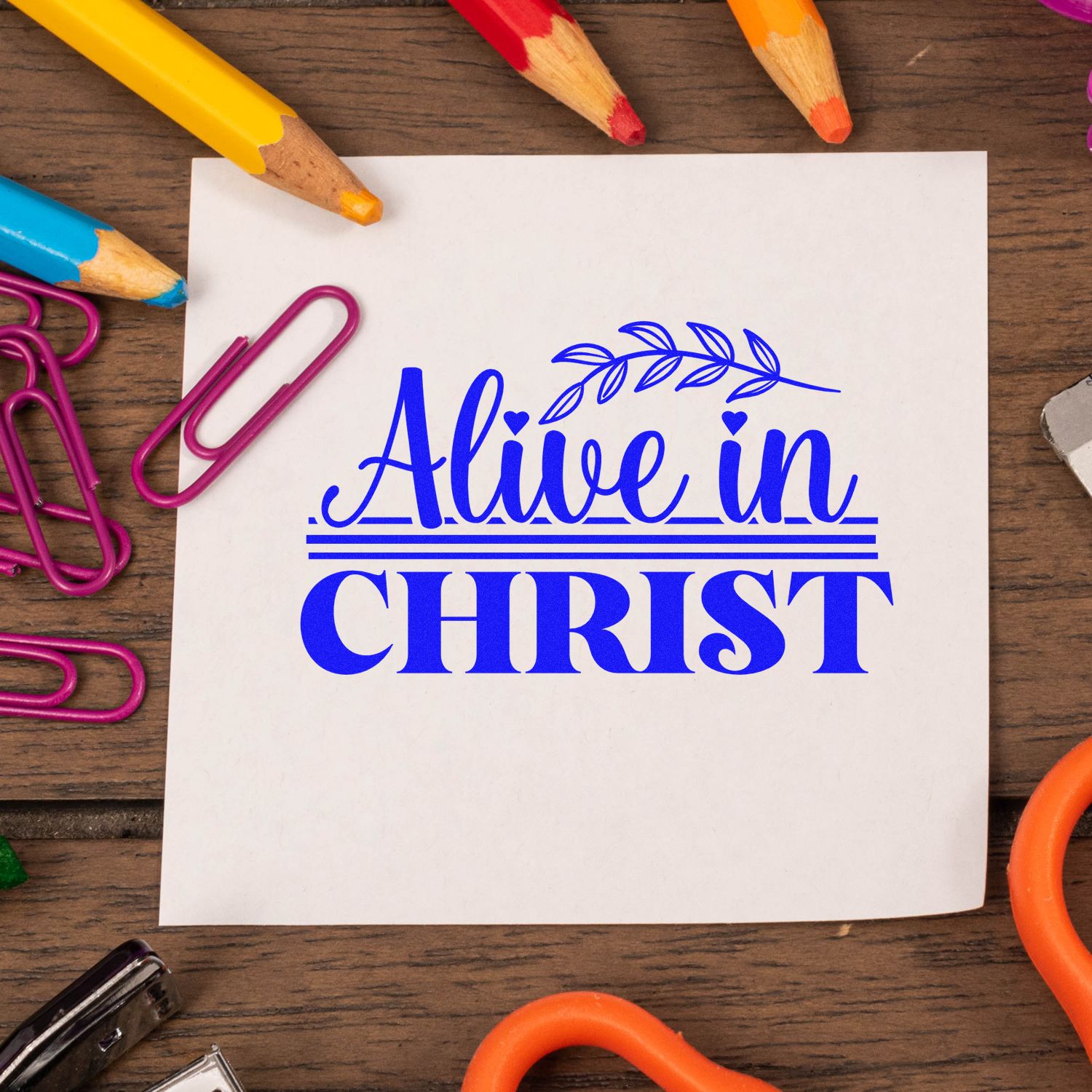 Blue Alive in Christ stamp and seal design on white paper, framed by colorful pencils and pink paper clips on wood.