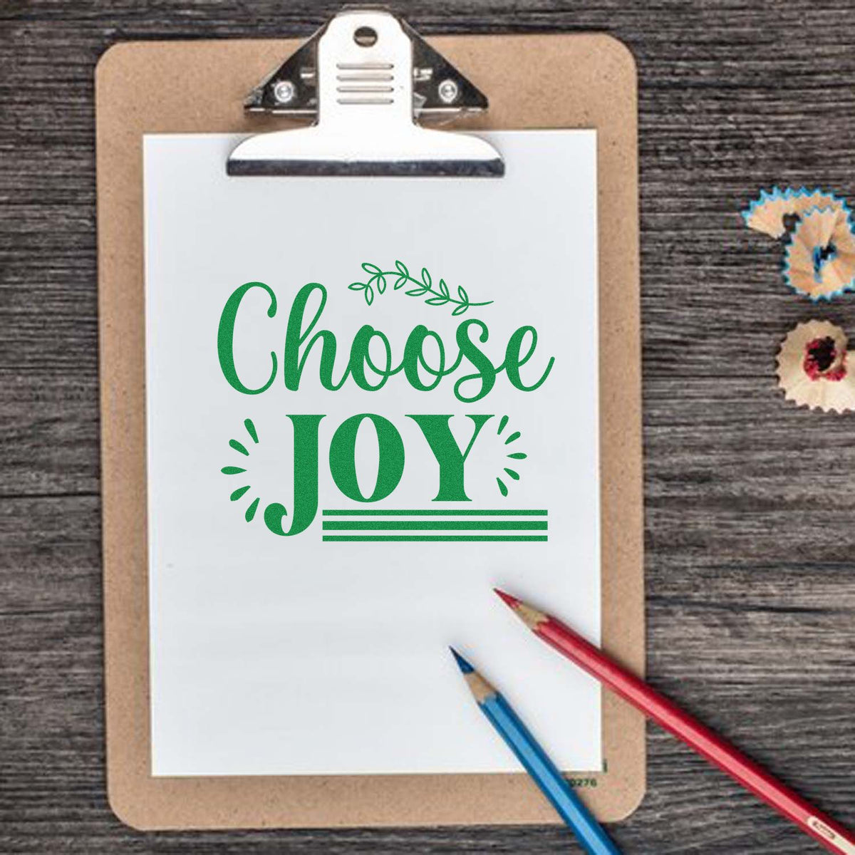 Green Choose Joy stamp and seal design on white paper clipboard, with pencils on a dark wood background.