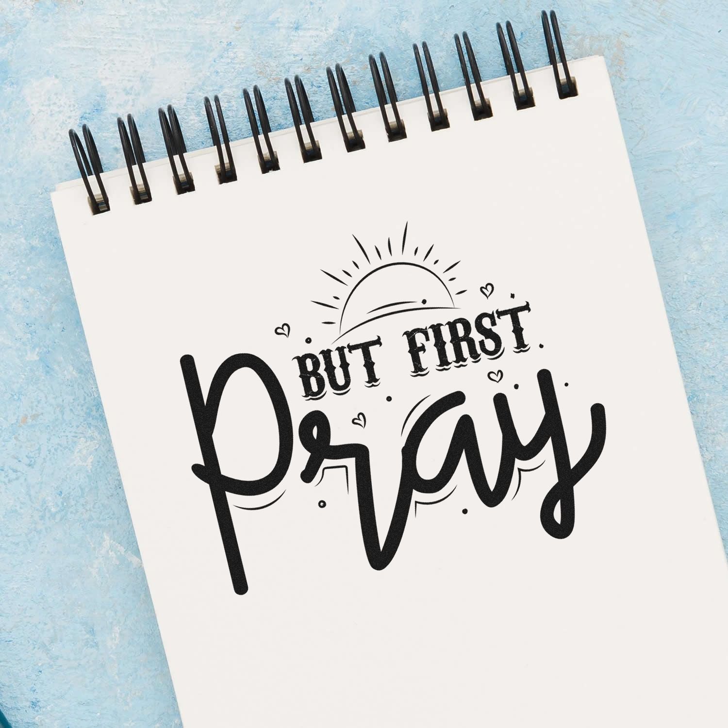 Black But First Pray stamp design on a white spiral notebook, with sun motif and hearts on a soft blue background.