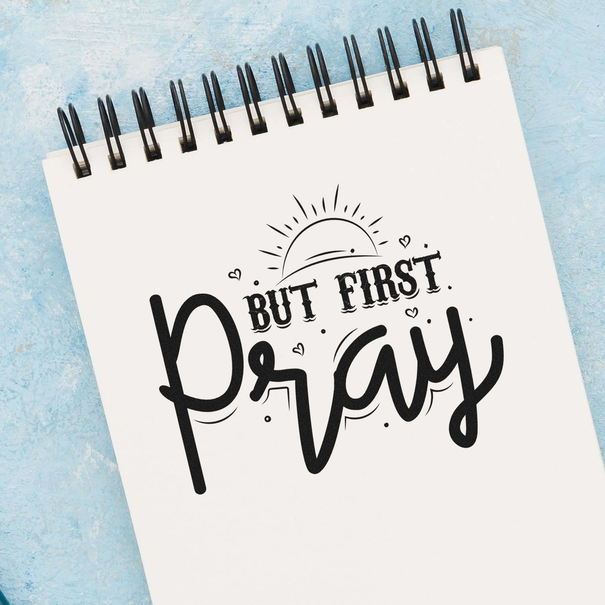 Black stamp/seal design reading But First Pray on a white spiral notebook, with sun rays, hearts, and a blue background.