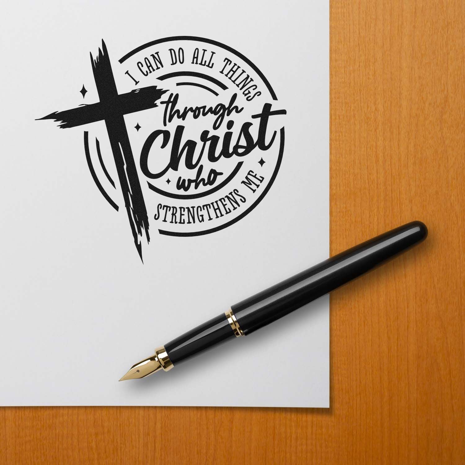 Black Christian stamp seal design with cross and quote on white paper, beside a black gold fountain pen on wood desk.