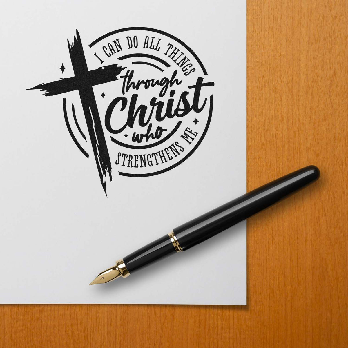 Black Christian stamp and seal design with cross and verse text on white paper, beside a black and gold fountain pen.