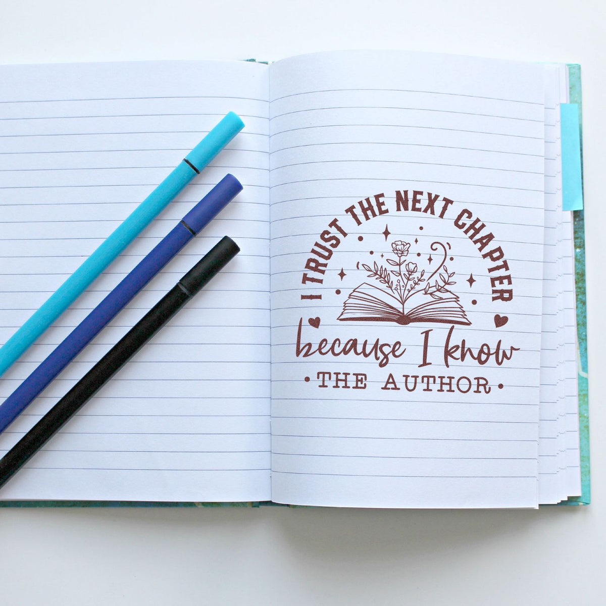 Brown book-lover rubber stamp on lined notebook with blue, purple and black pens on white pages.