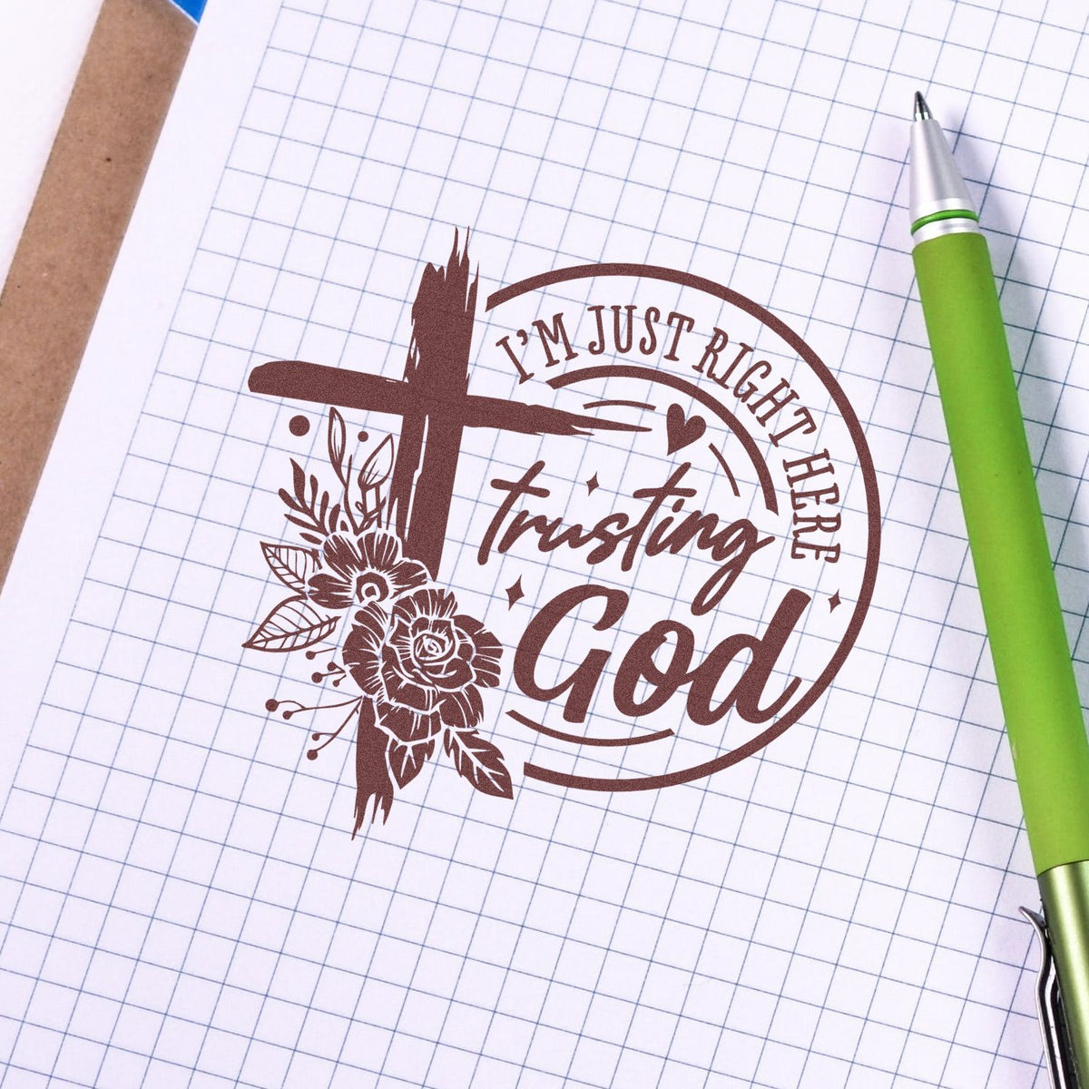 Brown custom stamp seal with floral cross and Im just right here trusting God on graph paper beside a green pen.
