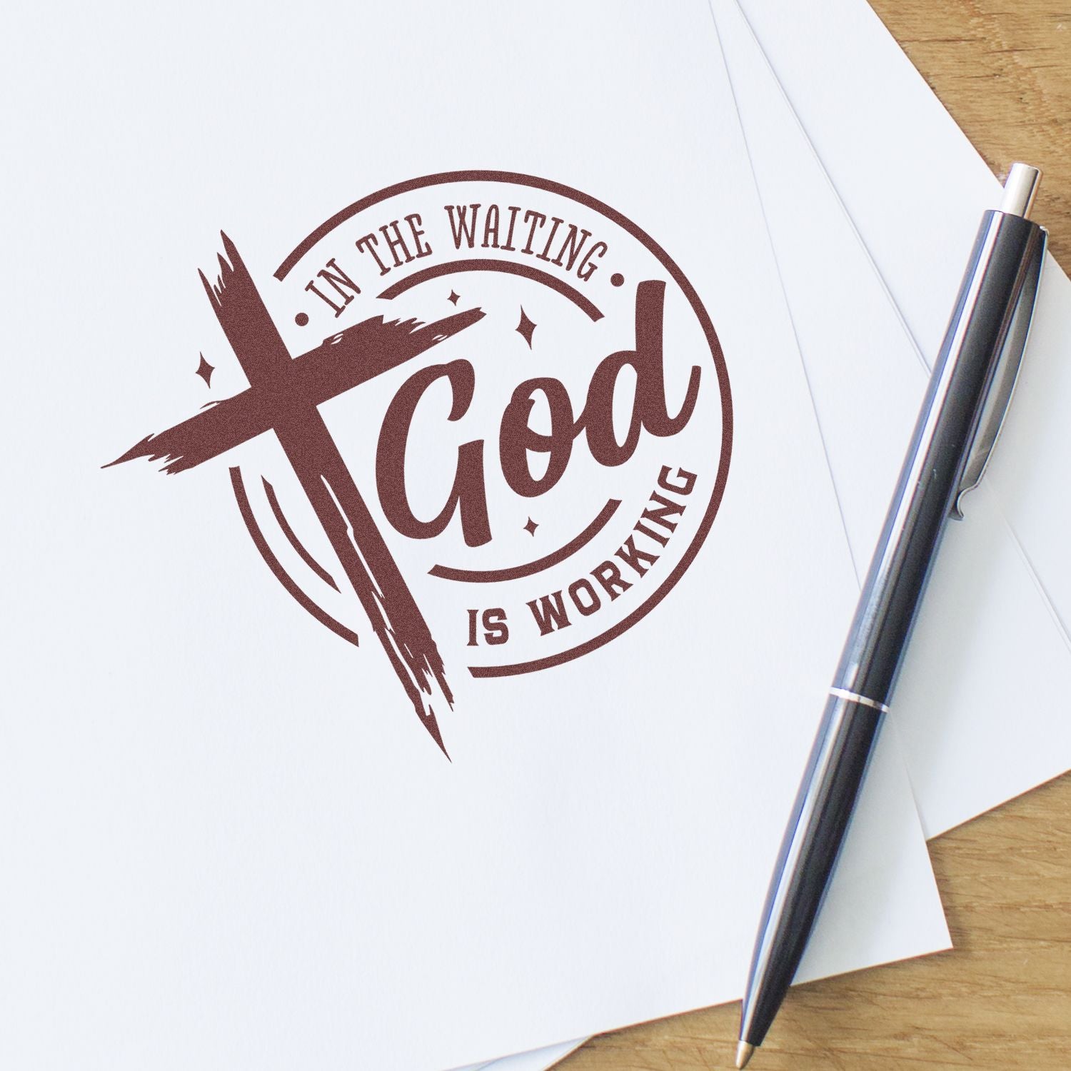 Brown custom rubber stamp seal reading In the waiting, God is working on white paper beside a black pen.