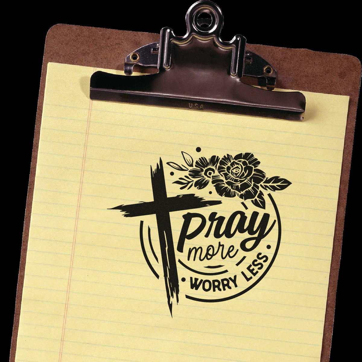Brown clipboard with yellow lined paper and a black rubber stamp seal reading Pray more, worry less with floral cross design.