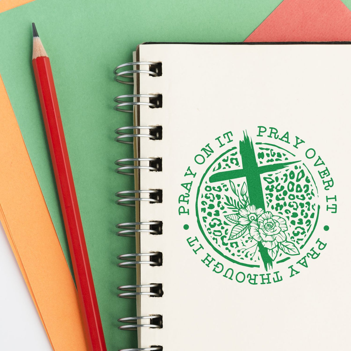 Green prayer floral stamp seal on a spiral notebook, beside a red pencil on pastel green, orange, and coral paper.