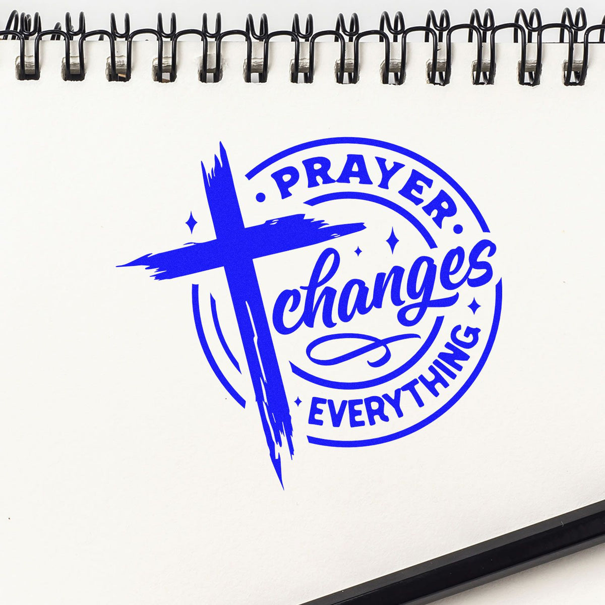 Blue rubber stamp seal reading Prayer Changes Everything with a cross on a white spiral notebook.
