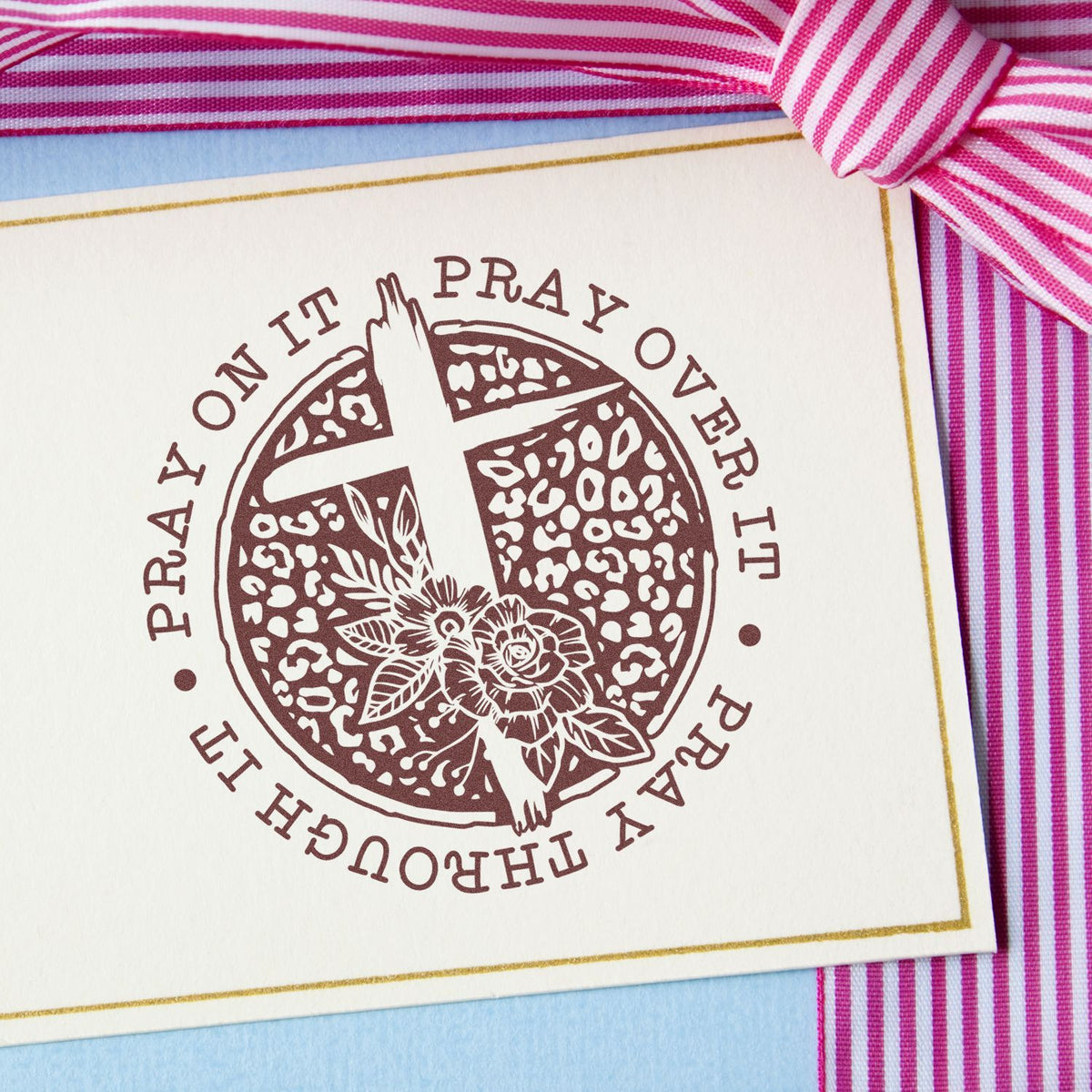 Brown prayer stamp seal with floral cross on cream card, gold border, pink striped ribbon, and blue background.