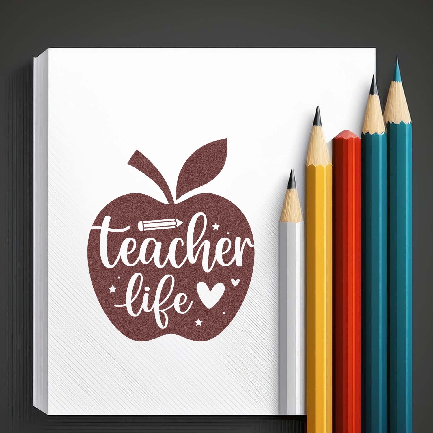 Brown teacher-life apple stamp seal on a white notebook, with yellow, red, teal and white pencils.