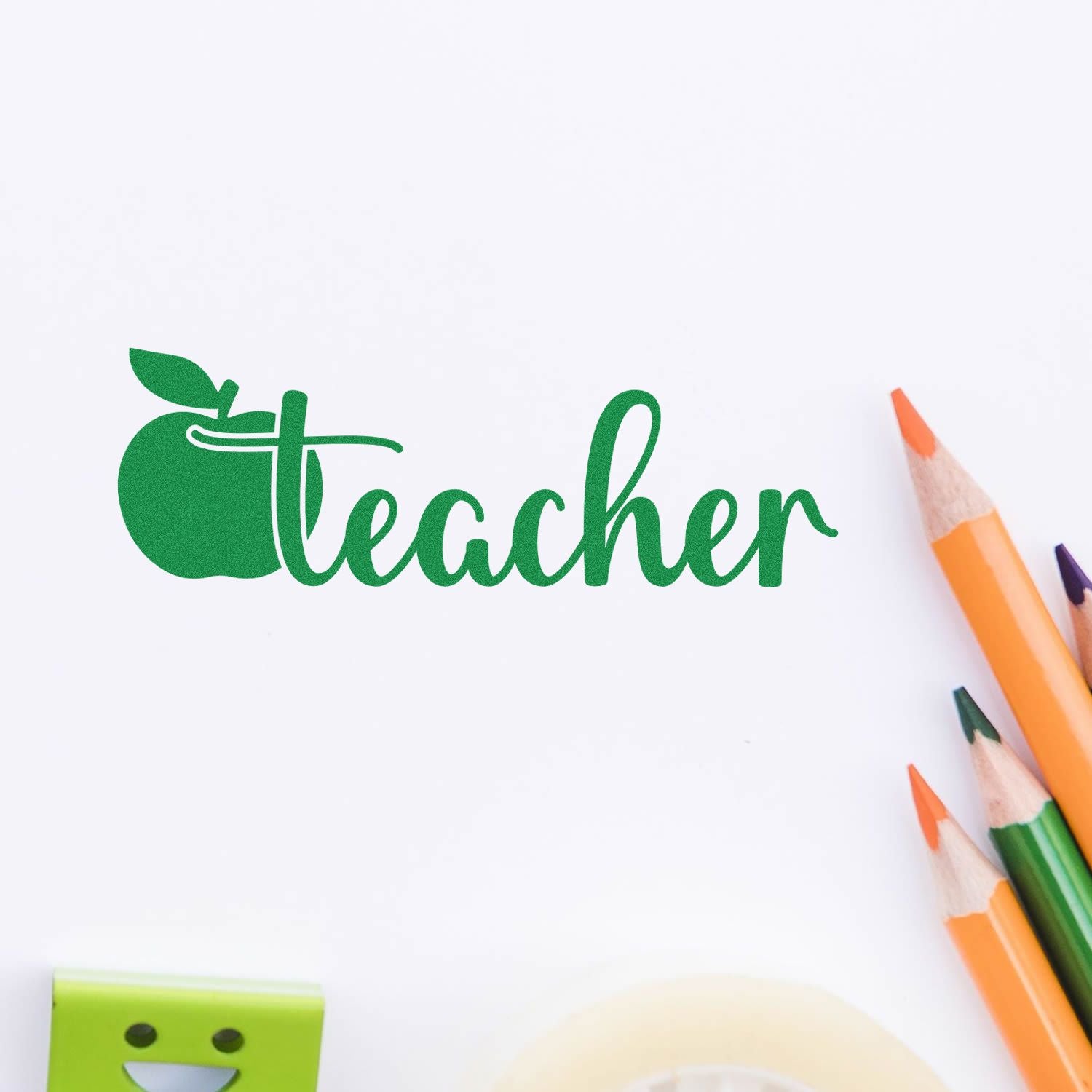 Green teacher stamp seal with apple icon on white paper, with orange and green colored pencils nearby.