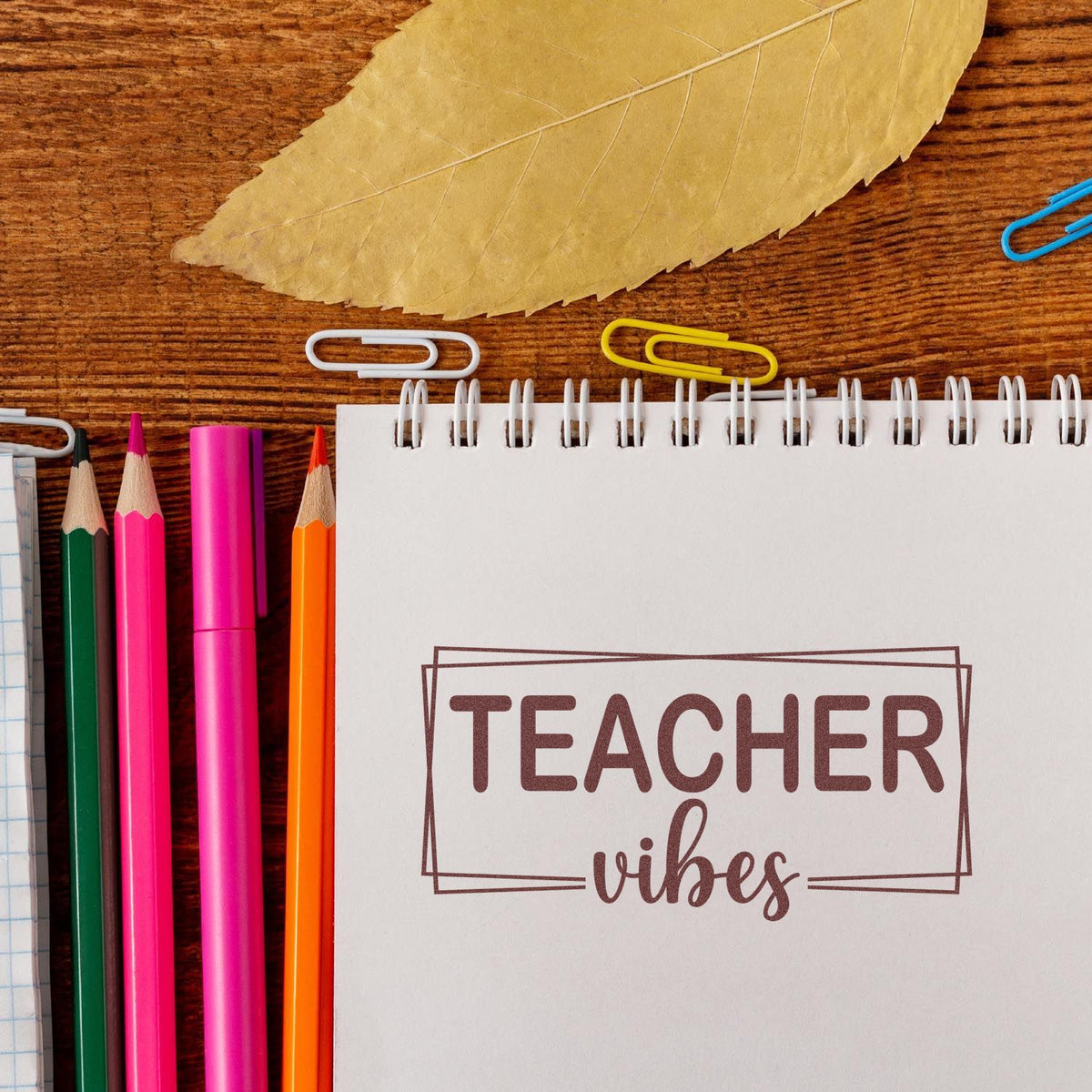 Teacher vibes stamp on white spiral notebook, with pink, green and orange pencils, paper clips, and a beige leaf on wood.