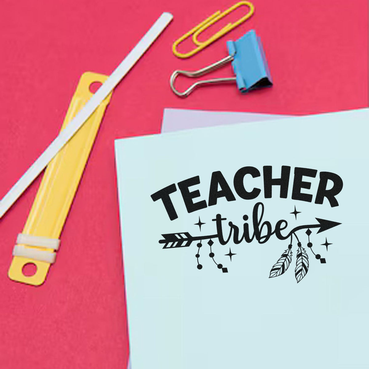 Teacher Tribe stamp seal in black on pale blue paper, with yellow and blue binder clips on a pink background.