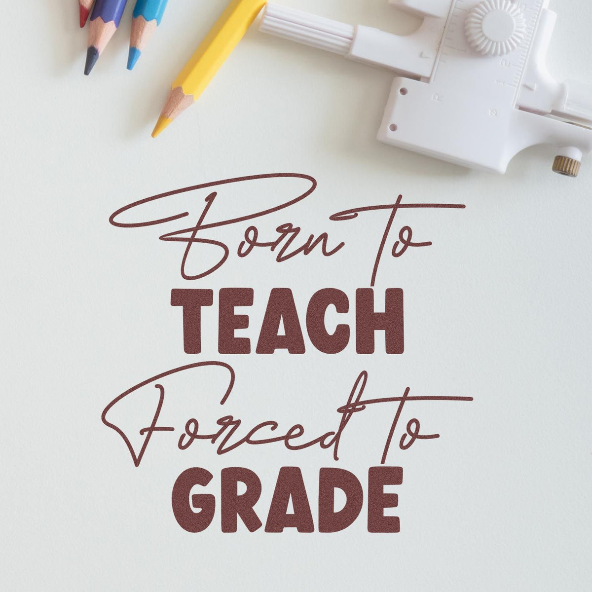 Maroon classroom stamp seal reading Born to Teach, Forced to GRADE, with colored pencils and white stamping tool.