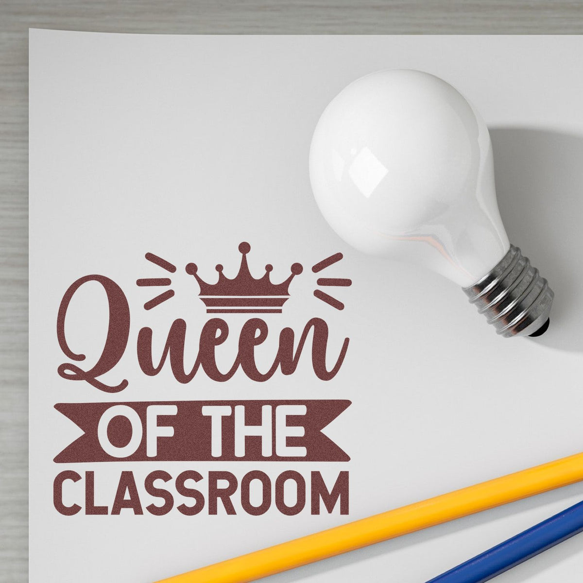 White light bulb beside maroon Queen of the Classroom custom stamp and seal design with crown on white paper.
