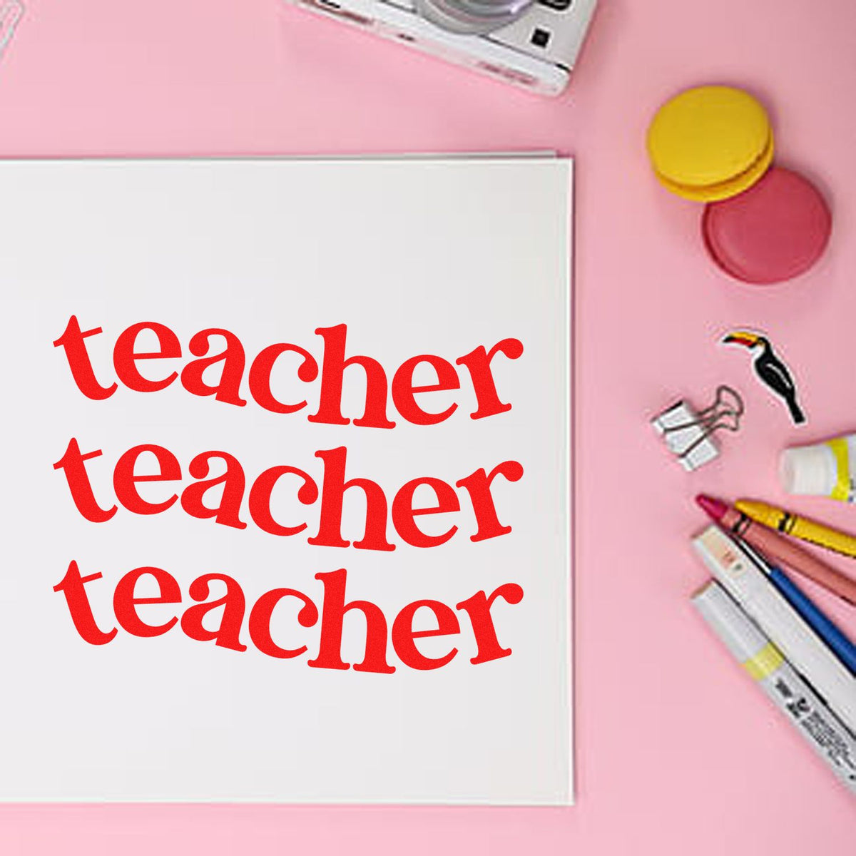 Red teacher stamp on white notebook, with pink background and colorful stamp and seal products, pens, and clips.