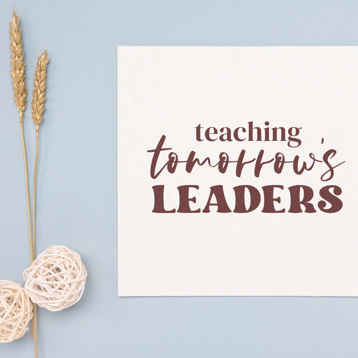 Maroon teaching, tomorrows leaders stamp/seal on white card, with dried wheat sprigs and white wicker accents on pale blue background.