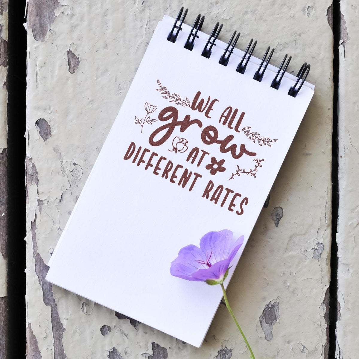 Brown floral stamp seal on white spiral notepad with we all grow at different rates text and purple flower.