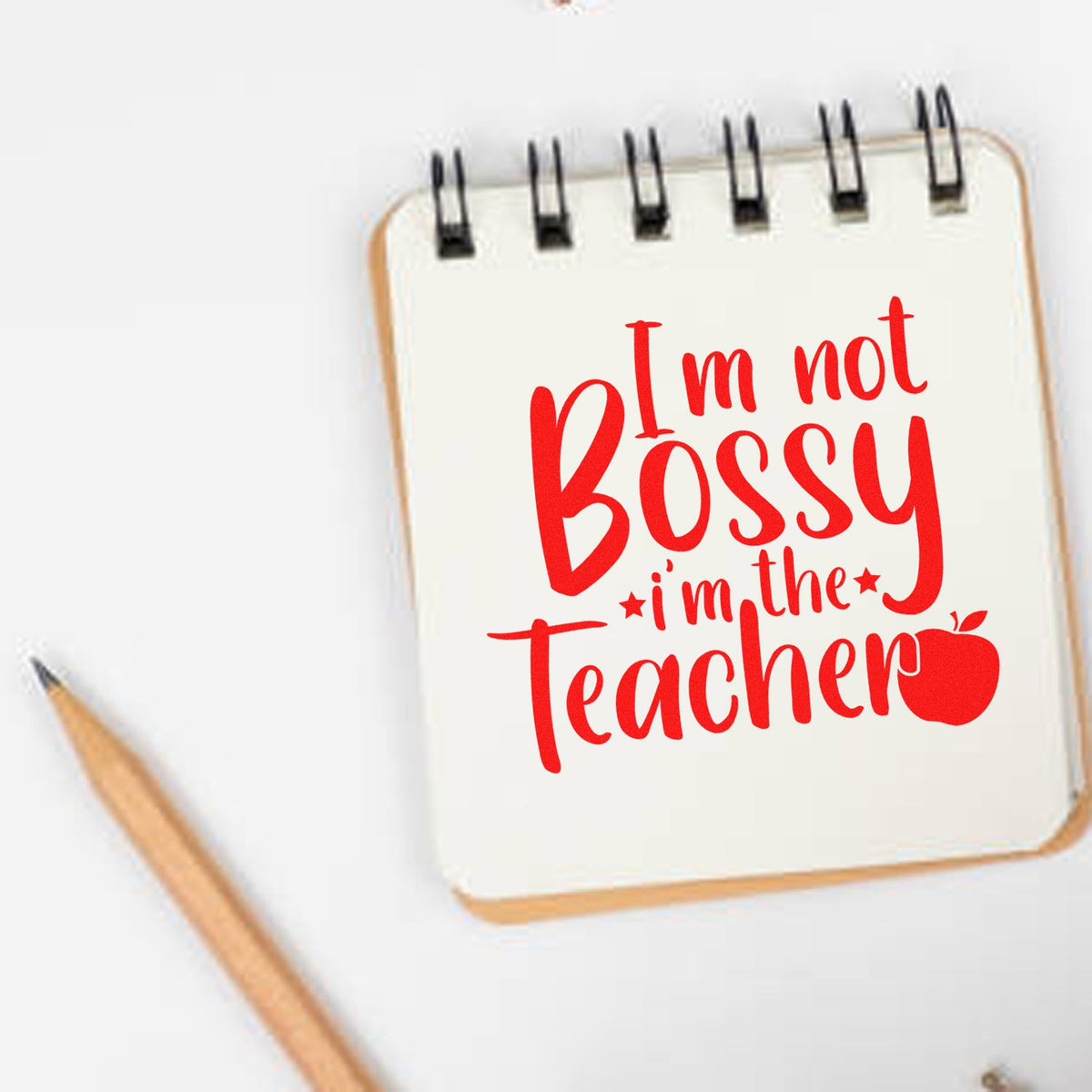 Red teacher stamp/seal design on white spiral notebook, reading "I'm not Bossy, I'm the Teacher," with pencil.