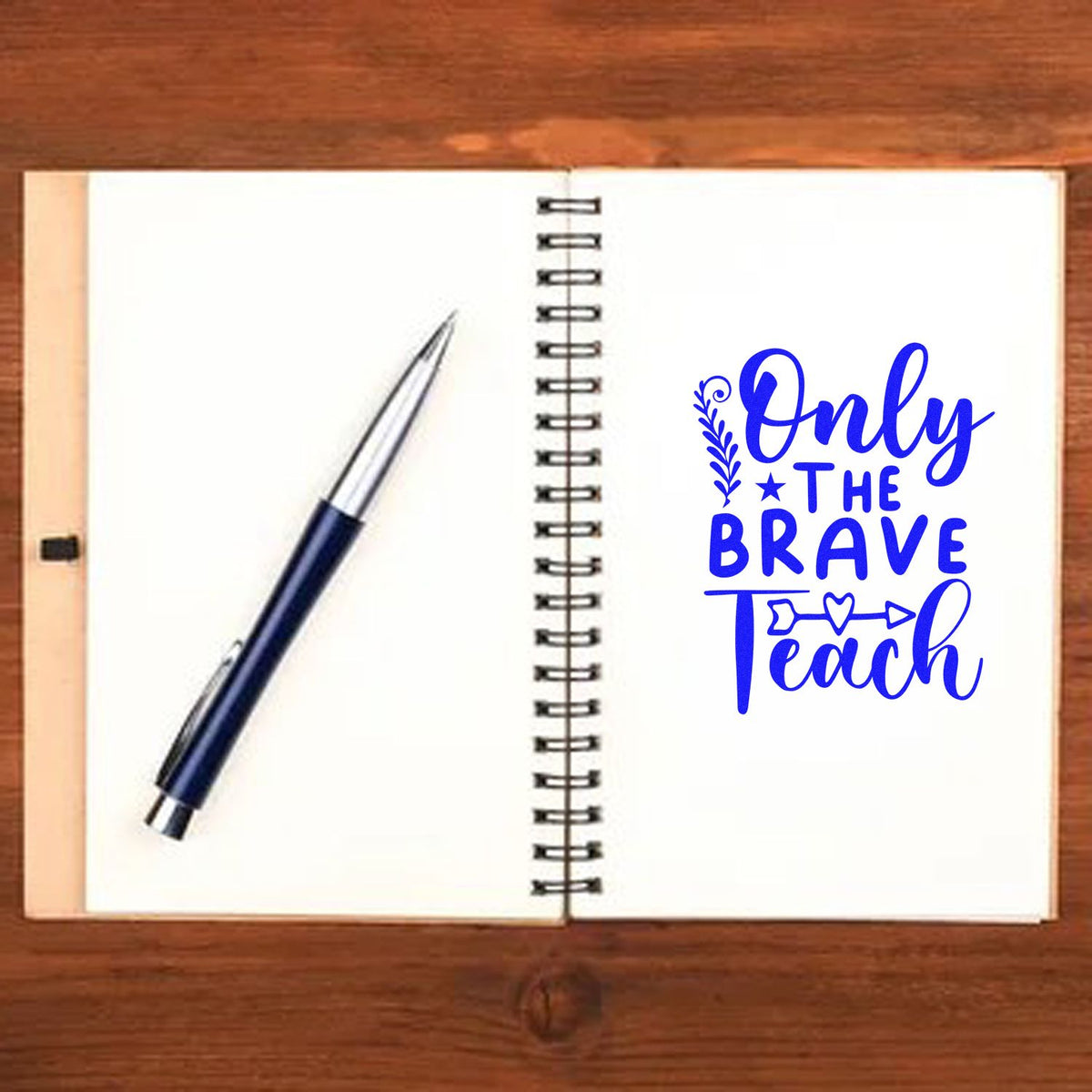 Blue stamp seal reading Only the Brave Teach on open spiral notebook beside a navy pen on a wooden desk.