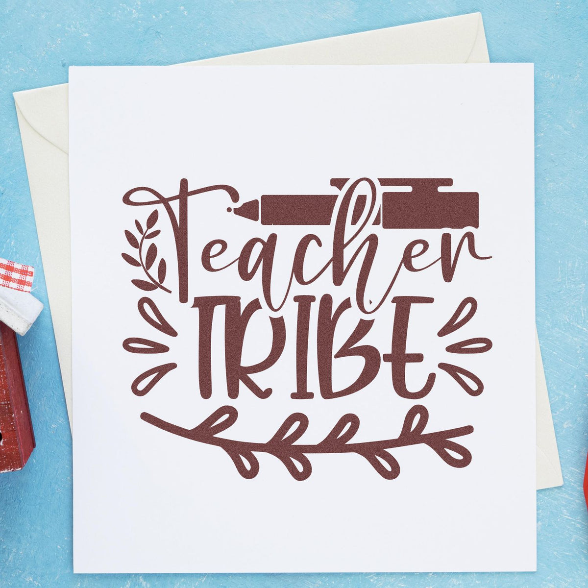 Brown Teacher Tribe rubber stamp and seal design with pen and leafy wreath on white card against a blue background.