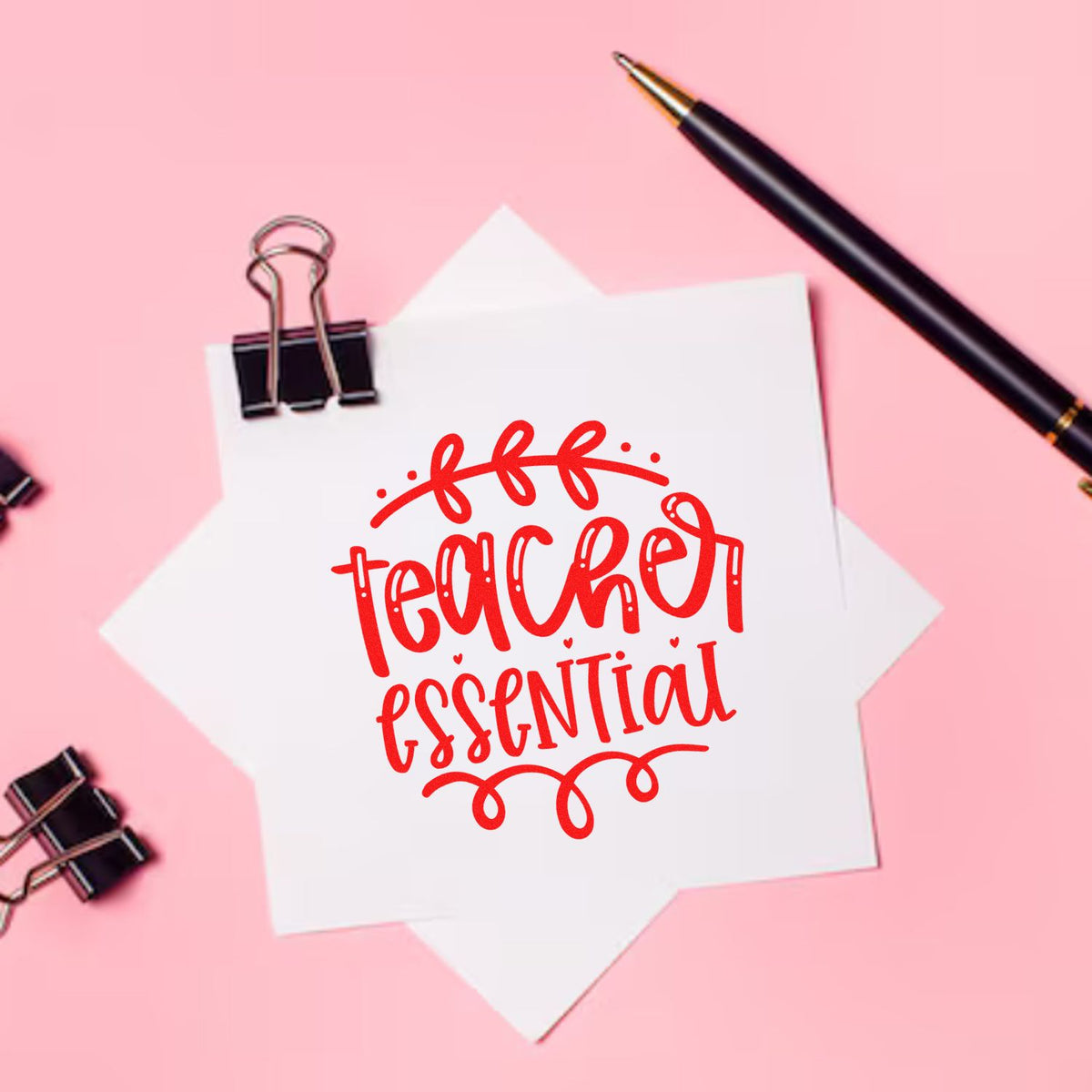 Pink stationery flat lay with white paper, red teacher essential stamp and seal design, black pen, and binder clips.