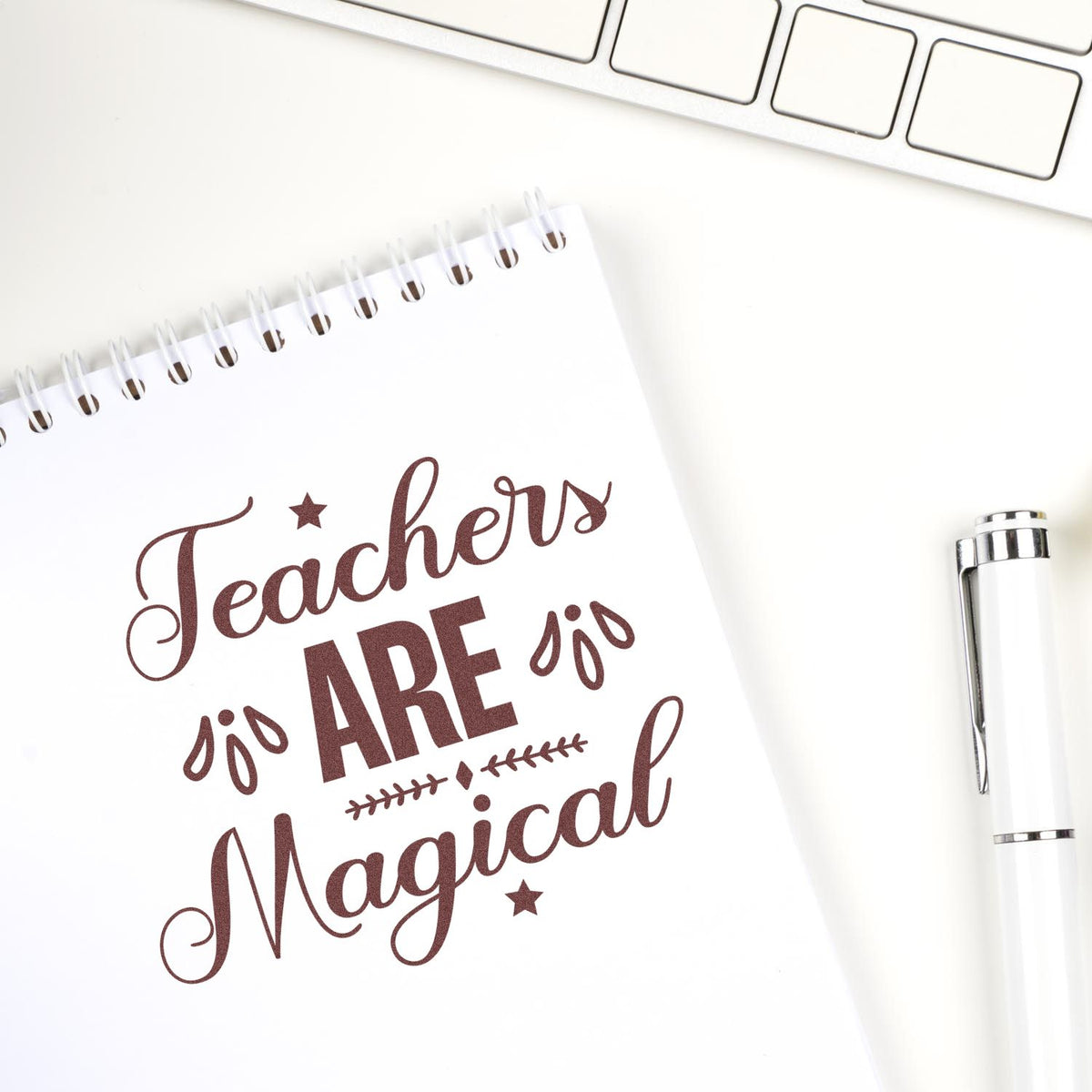 Brown Teachers are Magical stamp/seal on white spiral notebook, with silver pen and keyboard on a white desk.