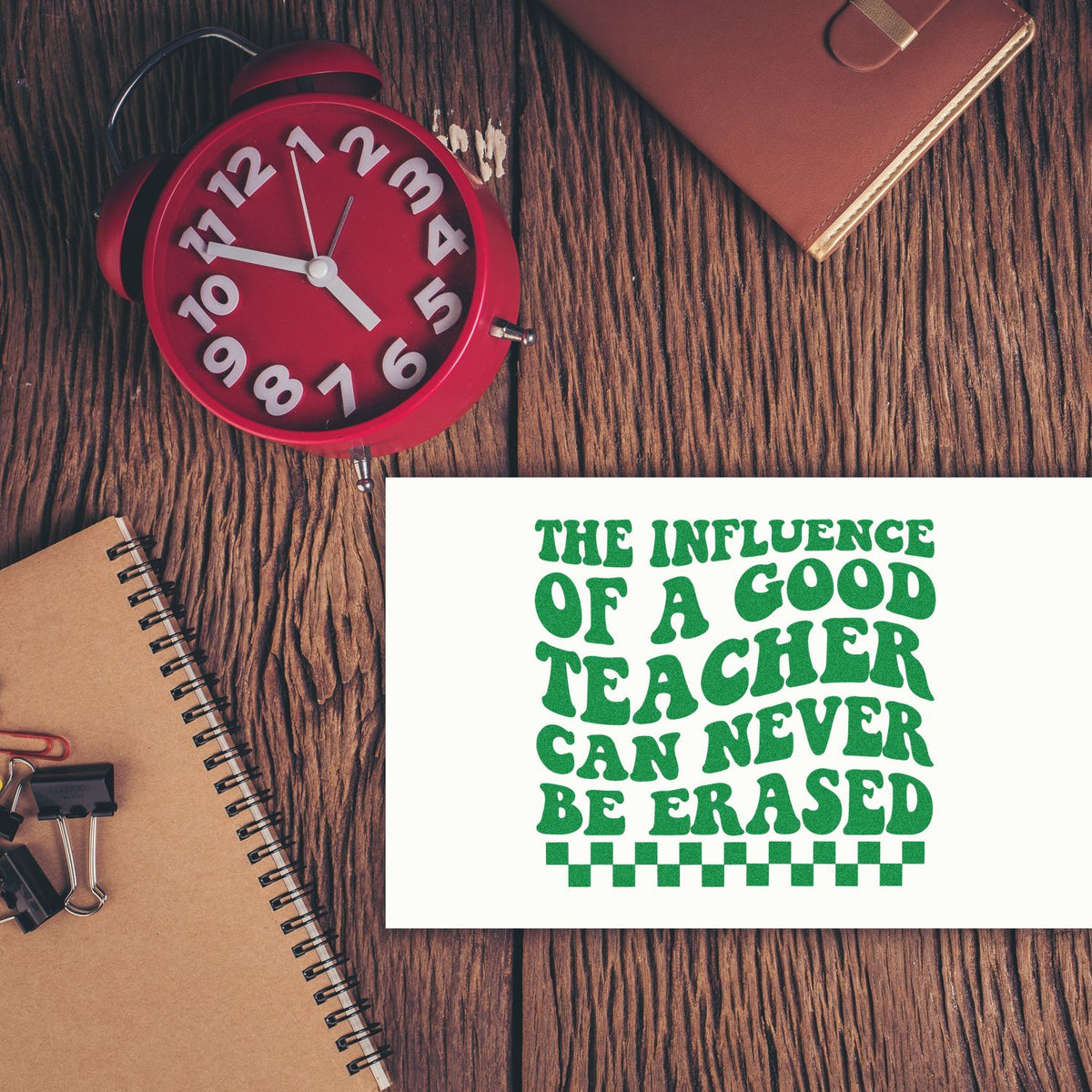 Green teacher quote stamp and seal on white card, with red alarm clock, brown notebook and wallet on a wood desk.