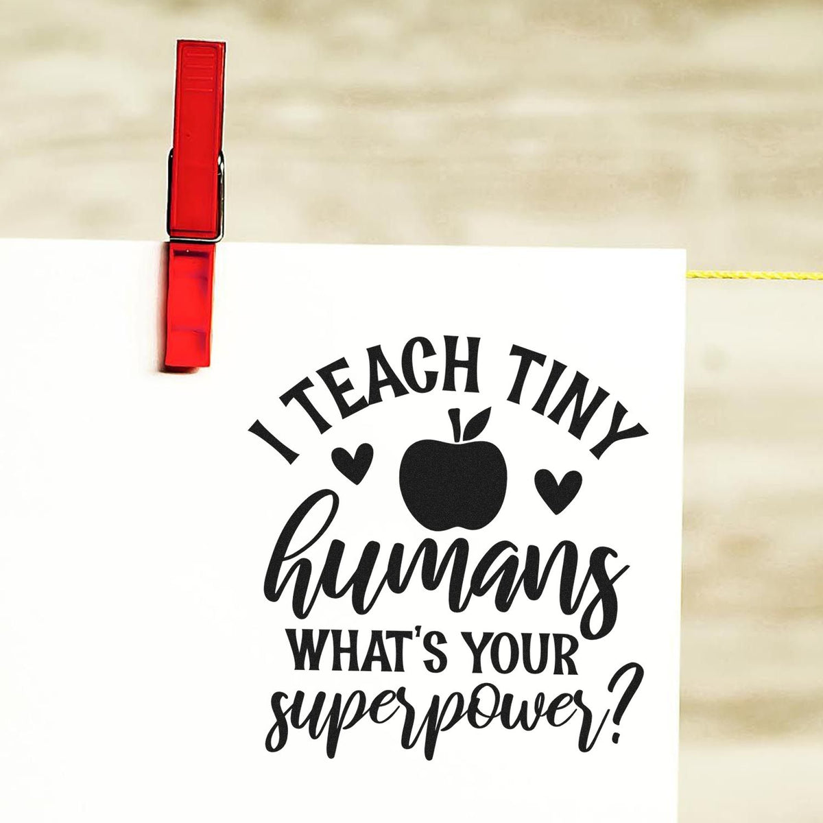 Red clip stamp seal holds a white card with black I teach tiny humans apple-and-hearts teacher design.