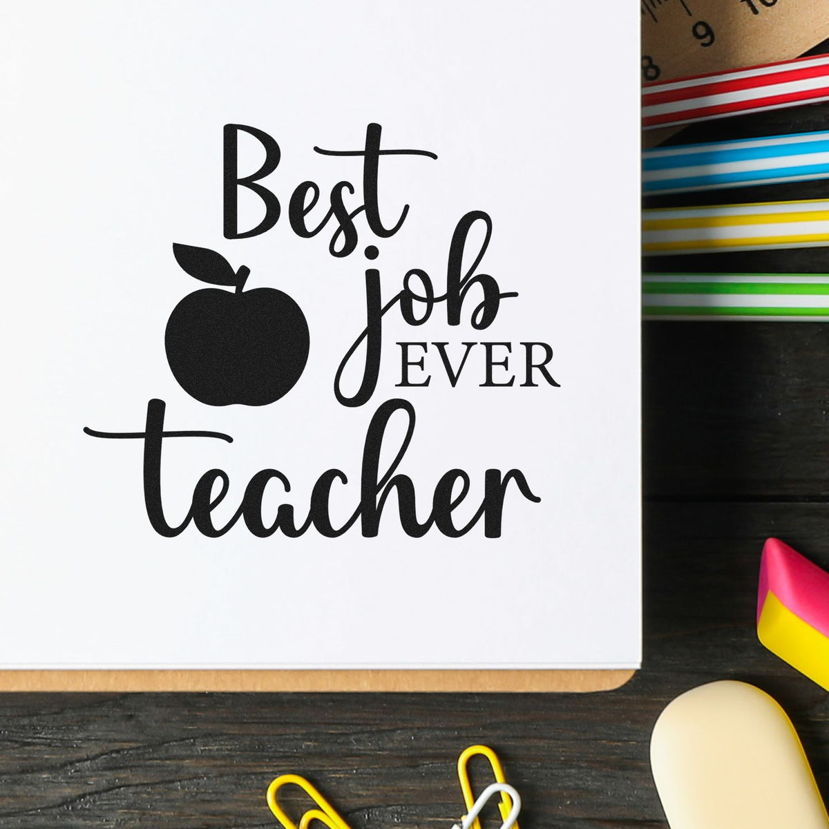 Black Best job ever teacher stamp with apple icon on white paper, beside colorful stamp pads and seal blocks on a dark desk.