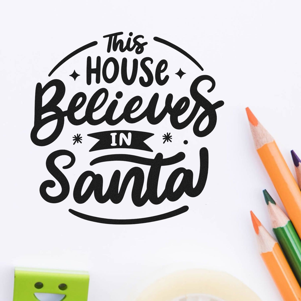 Black decorative house seal stamp reading This House Believes in Santa on white, with orange and green pencils nearby.