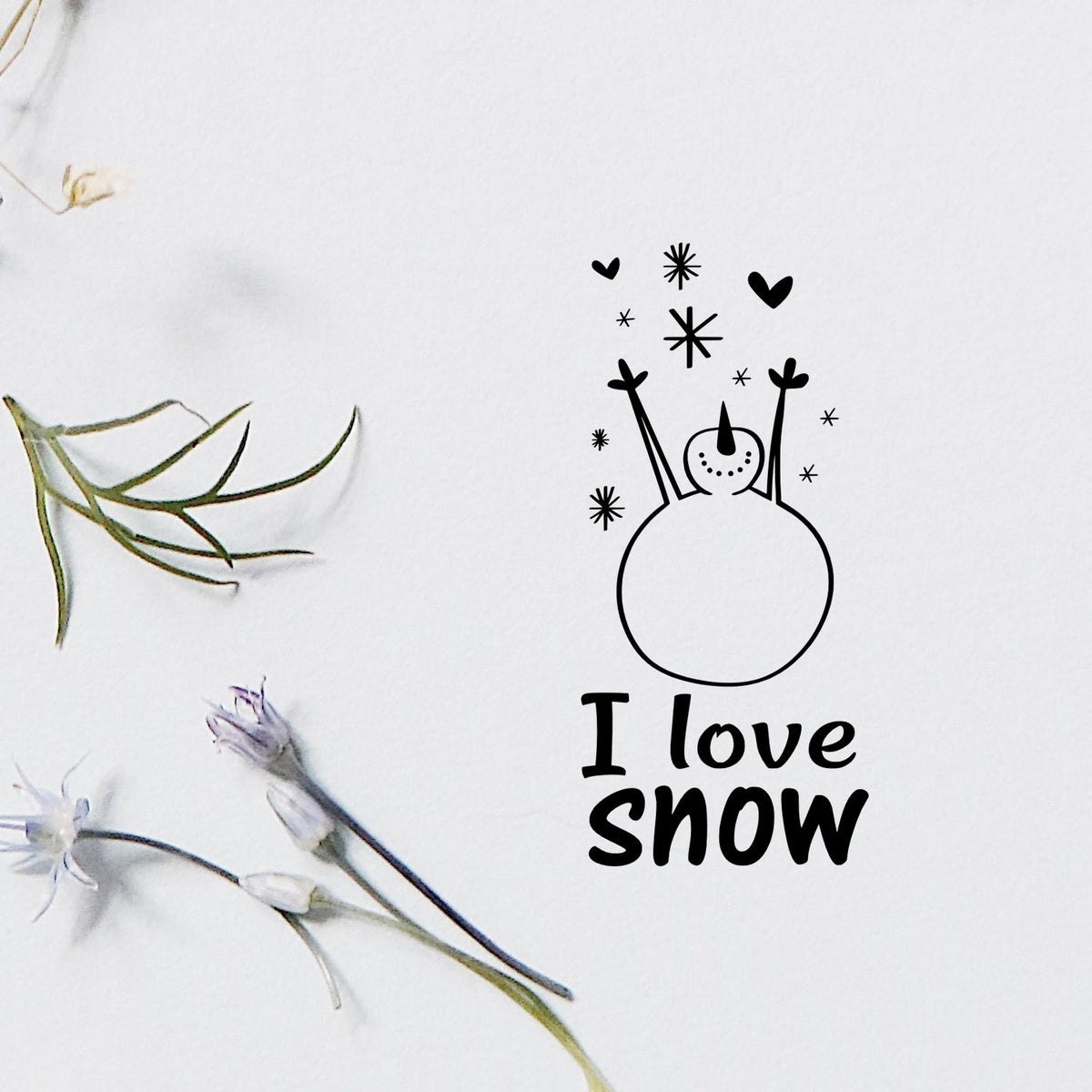 Black snowman stamp seal with hearts and snowflakes, reading I love snow, on white paper with dried flowers.