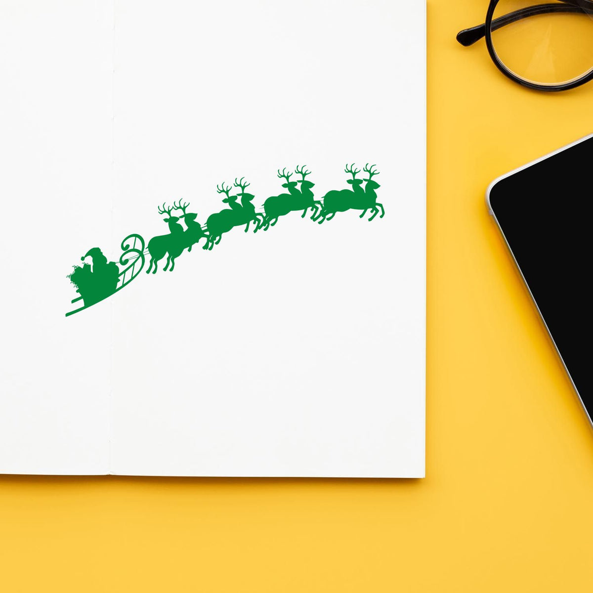 Green holiday stamp seal showing Santa in sleigh with reindeer on white paper, on yellow background beside black glasses and phone.
