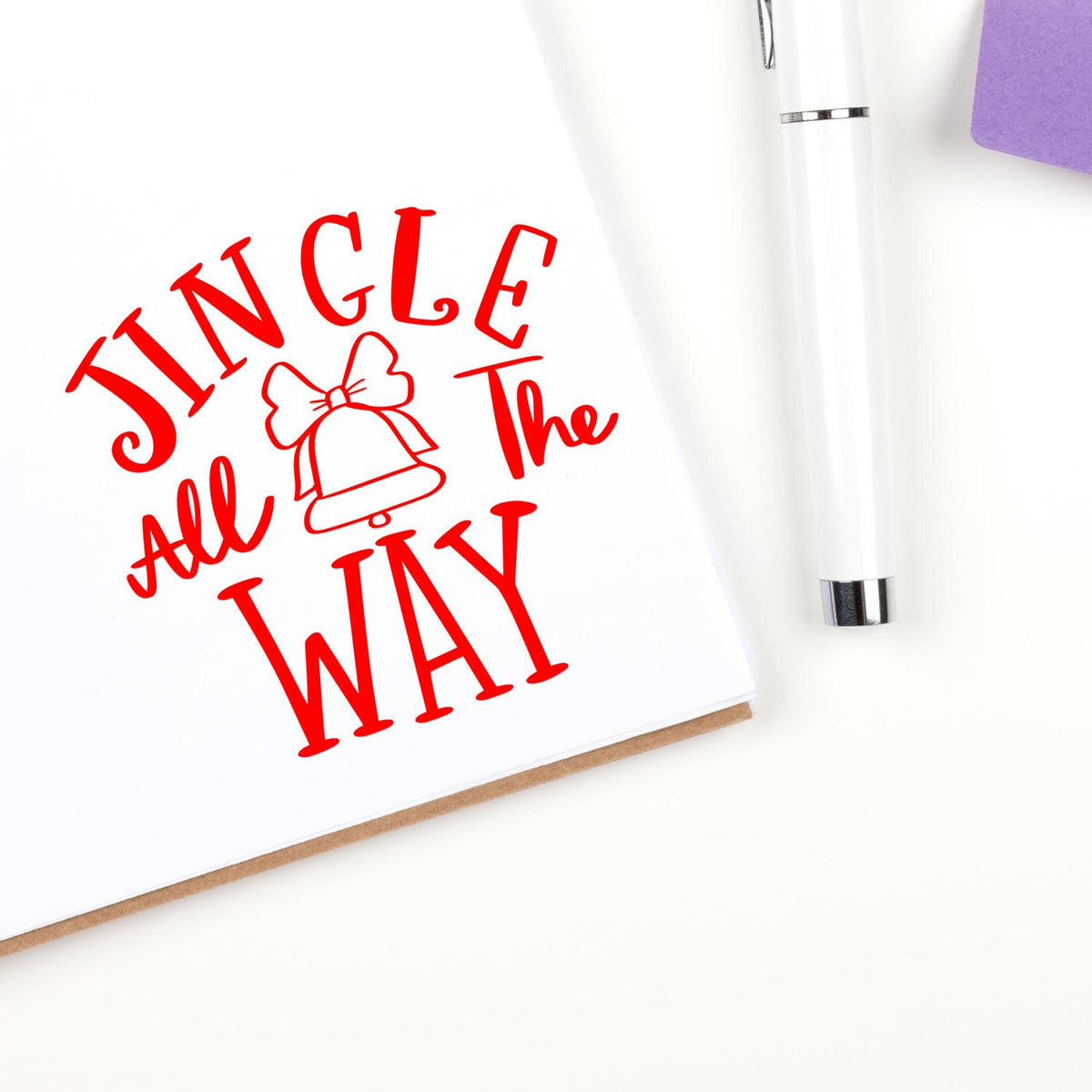 Red Jingle All the Way holiday stamp/seal with bell on white paper, beside a white pen and purple sticky note.