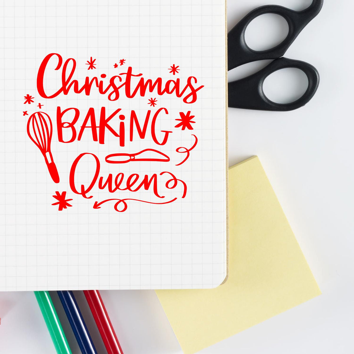 Red Christmas Baking Queen stamp design on grid paper, with black scissors, yellow note and colored pens.