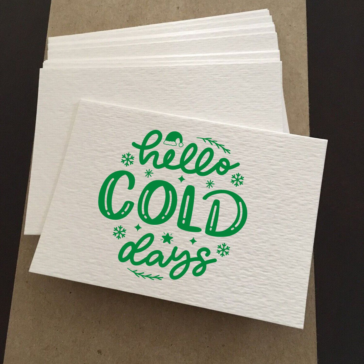 Stacked white stamp sheets with green hello cold days lettering, snowflakes, and winter sprigs for holiday seals.