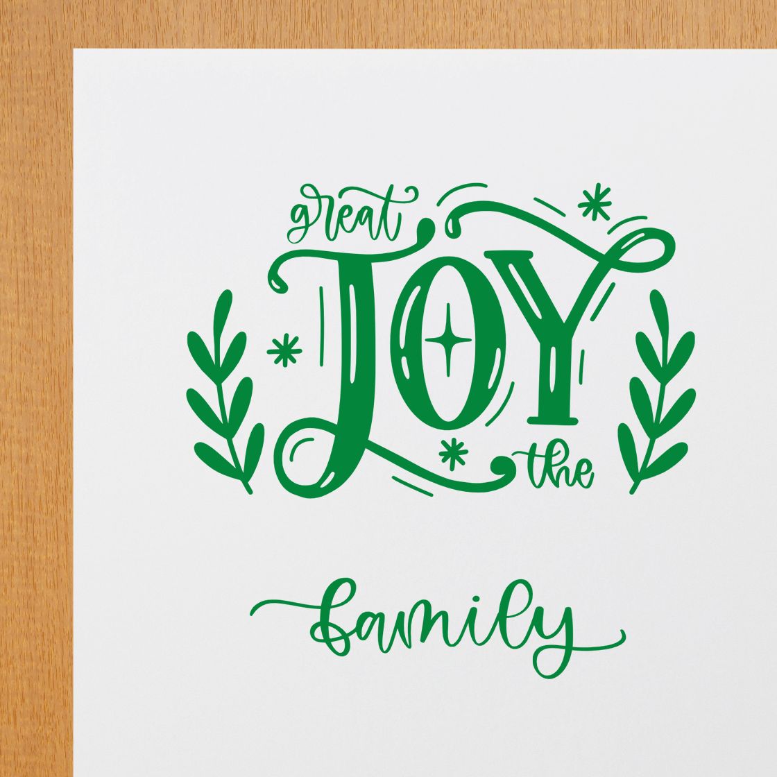 Green holiday stamp/seal design with Great Joy the Family typography, leafy accents, and starbursts on white paper.