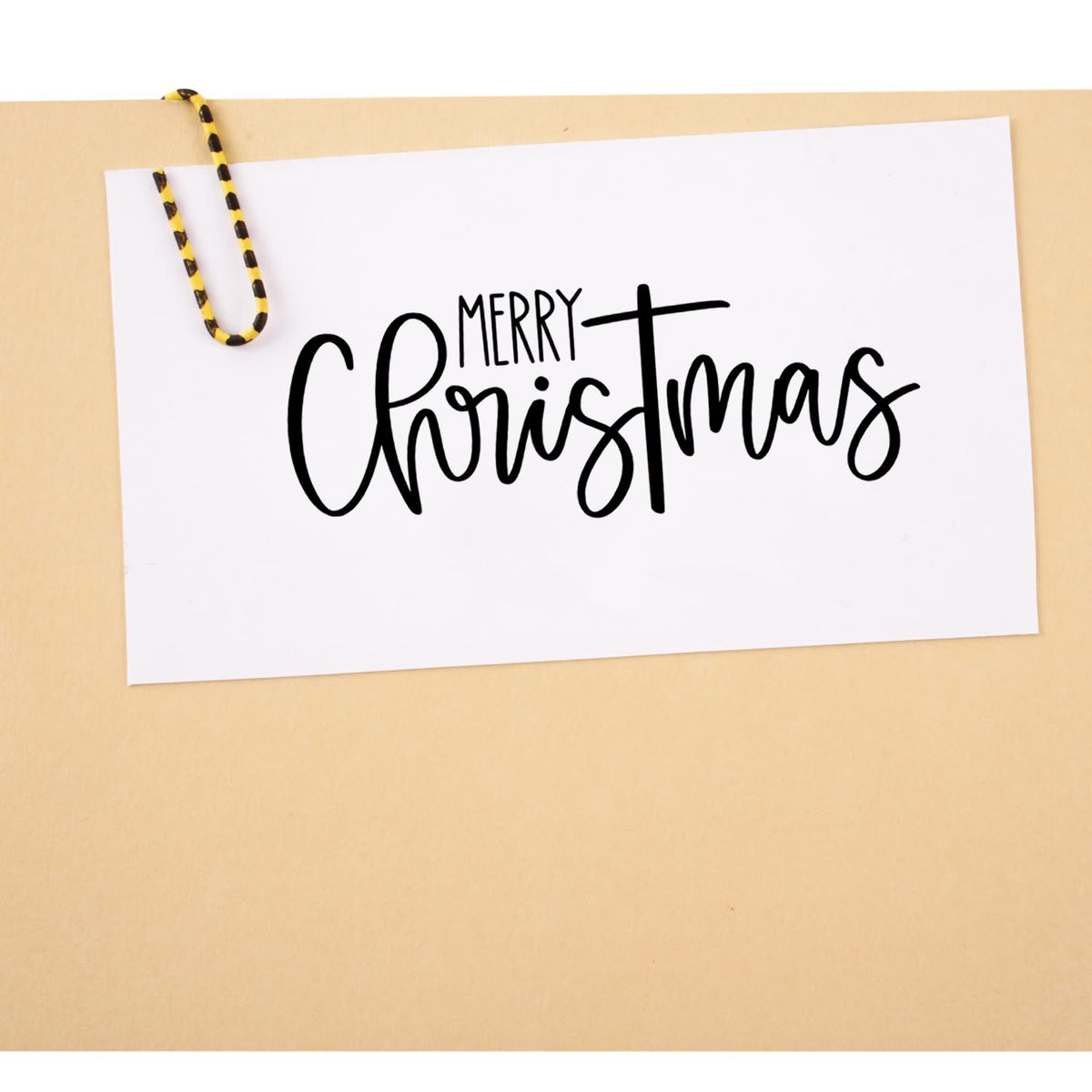 Black Merry Christmas stamp/seal on a white card, clipped with a yellow-and-black striped paperclip on a beige background.
