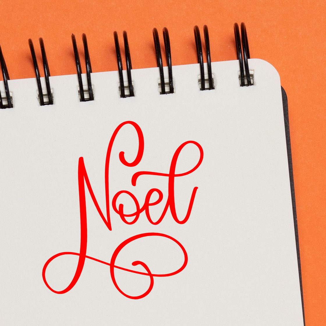 Red Noel script stamp/seal on a white spiral notebook with black rings against an orange background.