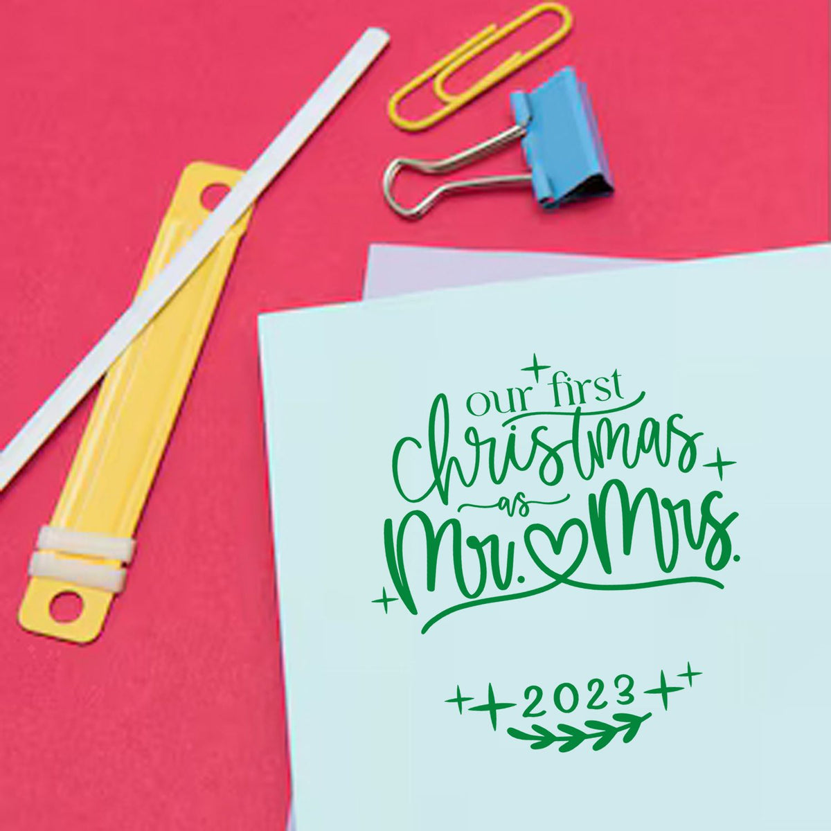 Green Our First Christmas as Mr. & Mrs. 2023 stamp on pale blue card, with yellow and blue binder clips on pink background.