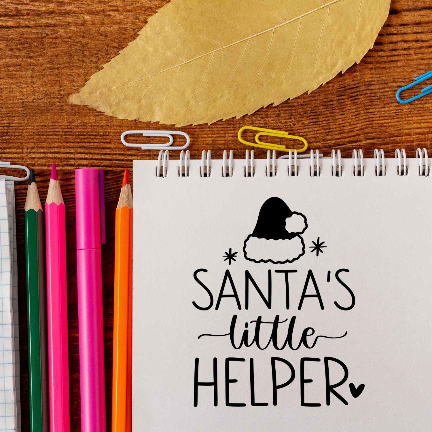 Holiday stamp and seal design reading Santas Little Helper on a white spiral notepad, with pink, green, and orange pencils.