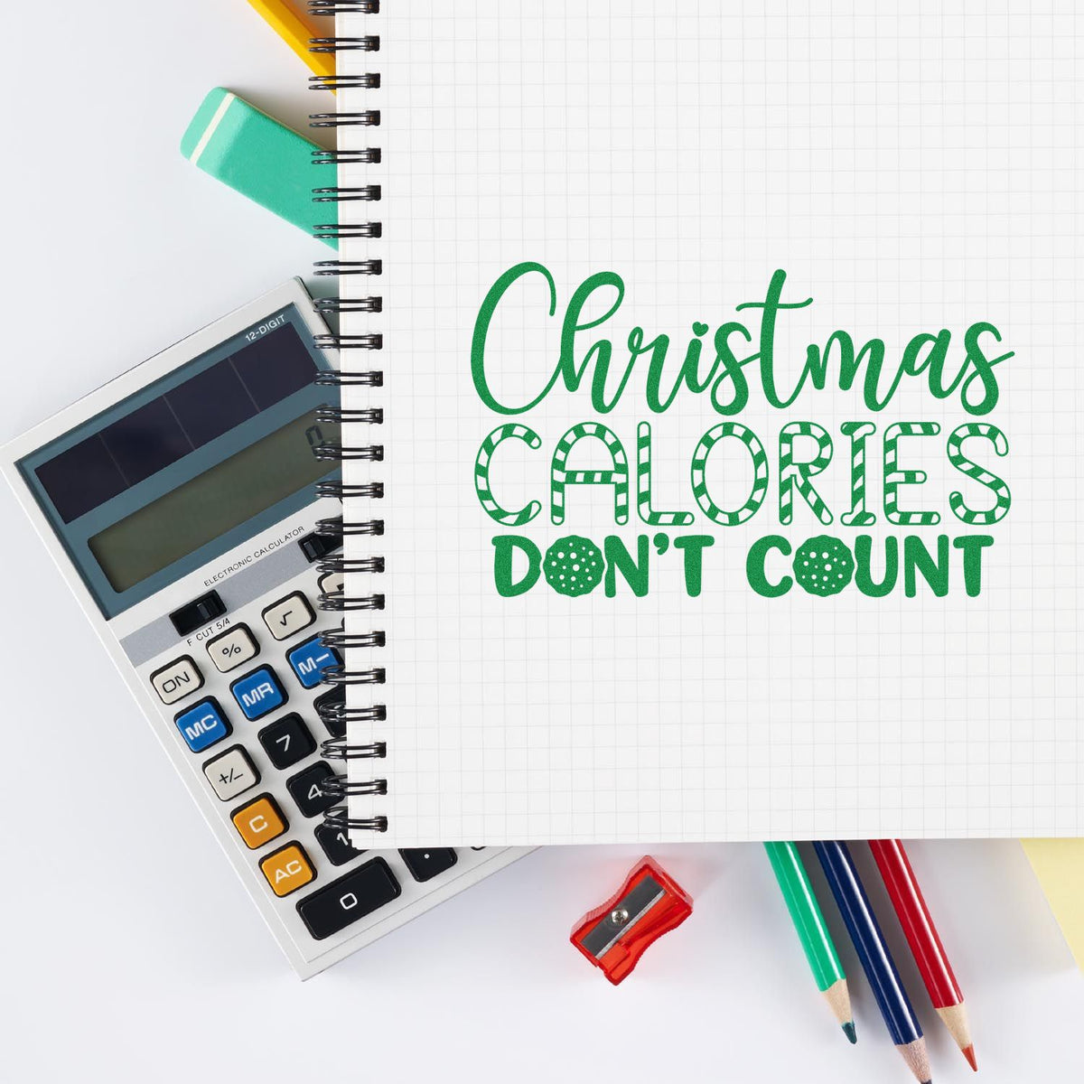 Green Christmas stamp and seal design on graph paper notebook, with calculator, pencils, and sharpener on white.