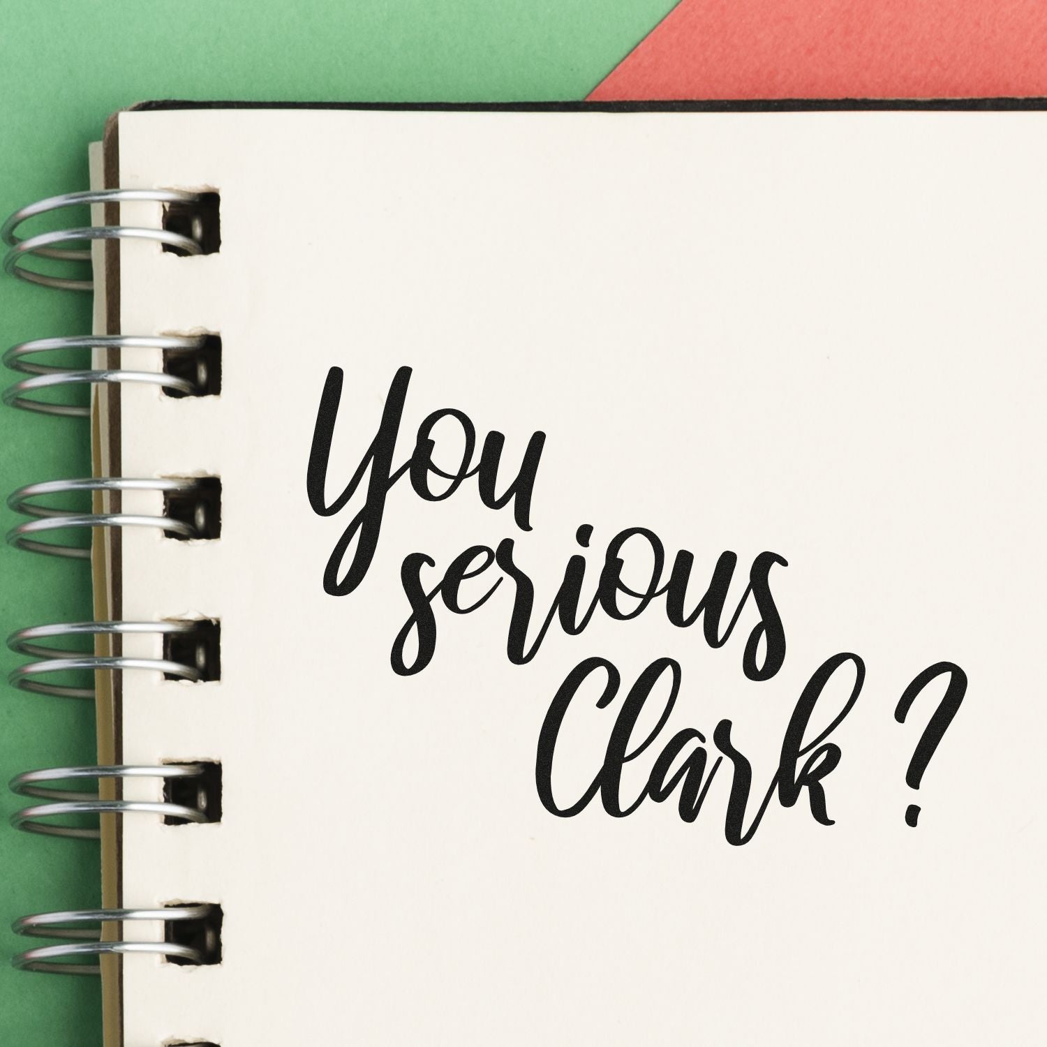 Black rubber stamp imprint on white spiral notebook page reading You serious, Clark? over green and red background.
