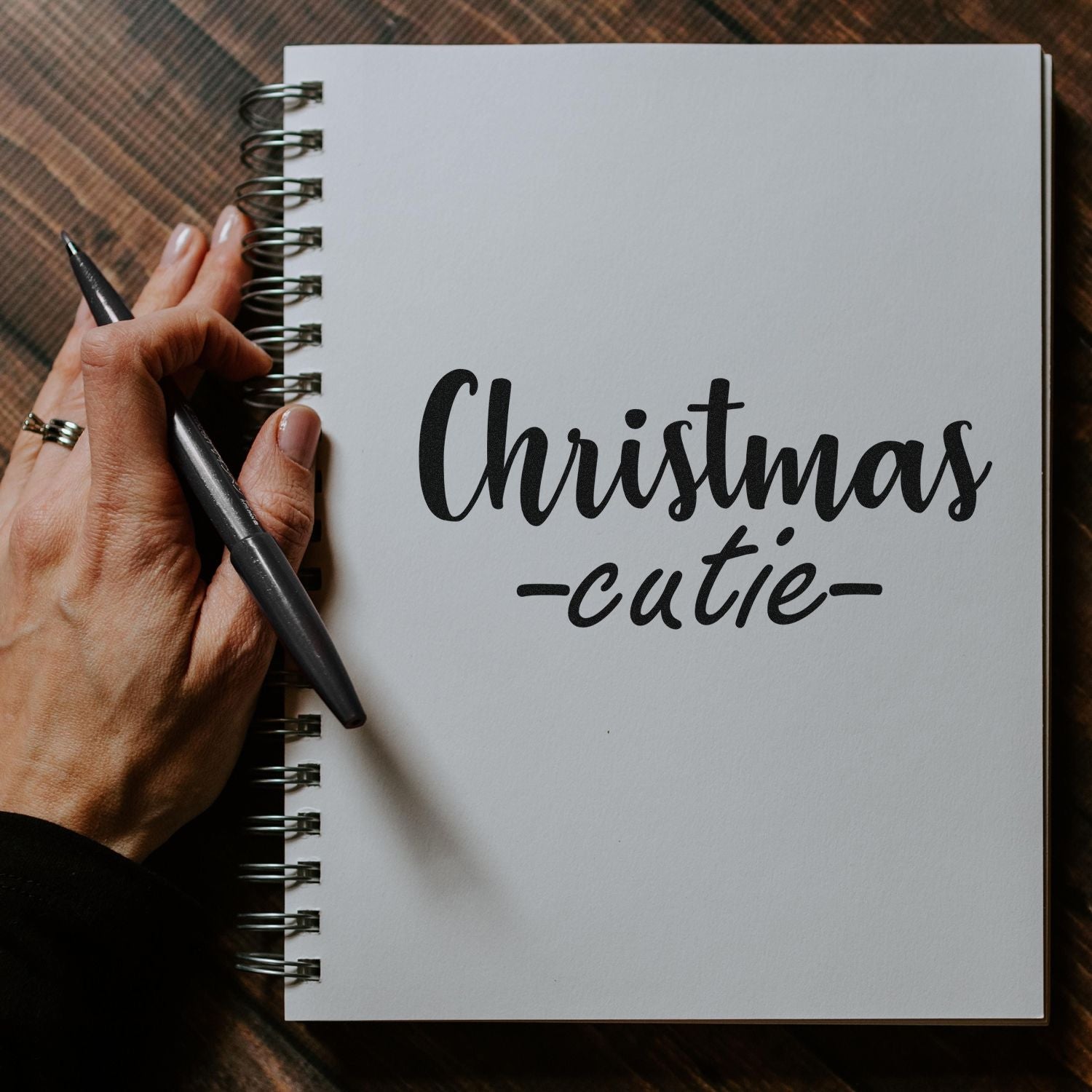 Black Christmas-cutie script stamp/seal on a white spiral notebook, with a hand holding a pen.