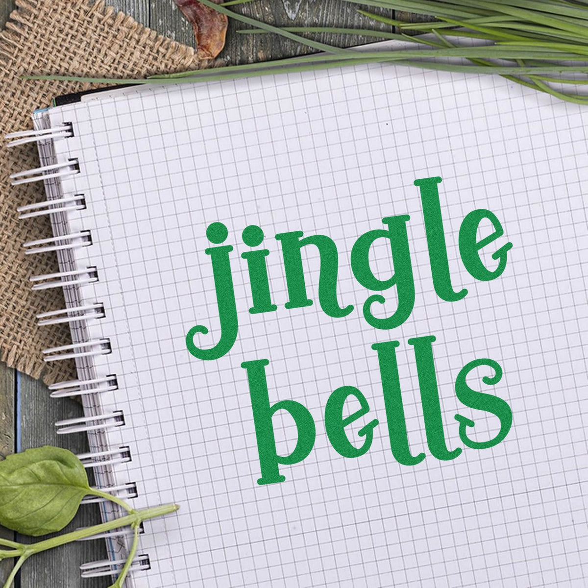 Green jingle bells stamp design on graph paper notebook, with spiral binding and rustic leaves for seal products.