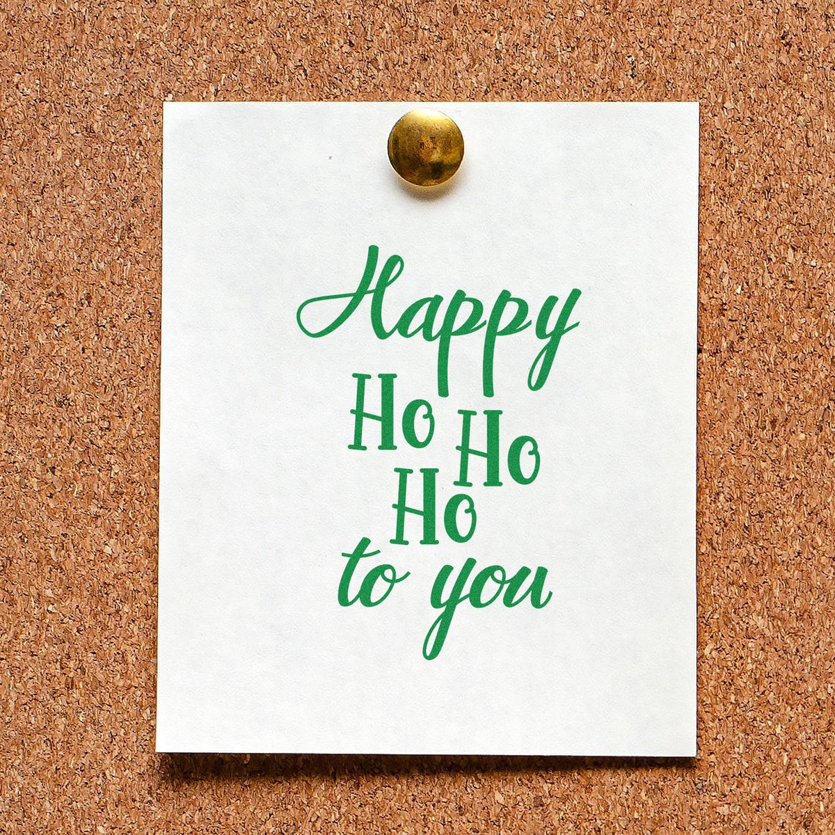 Green holiday stamp on white card with brass seal pin, reading Happy Ho Ho Ho to you on corkboard.