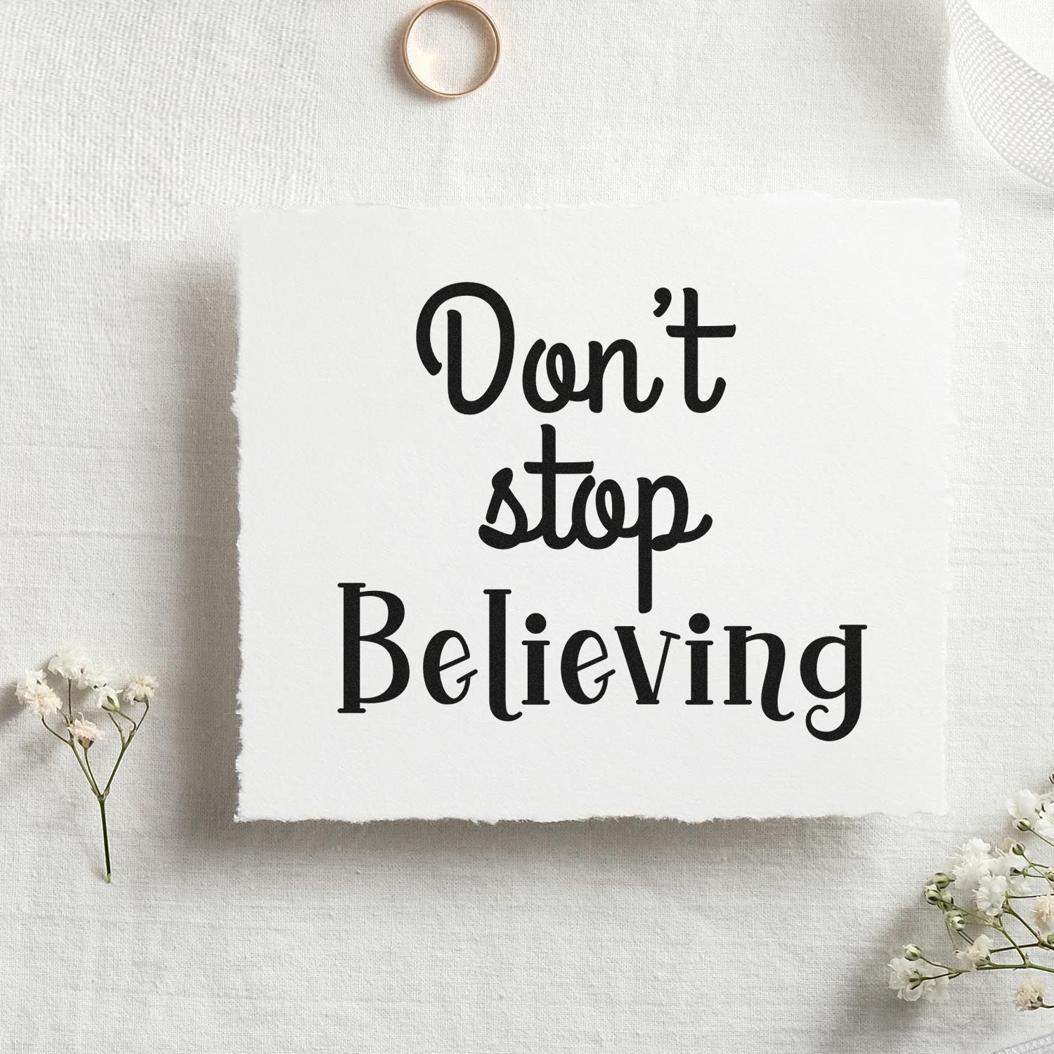Black Dont stop Believing stamp on white textured paper with minimalist seal-style design and delicate white flowers.
