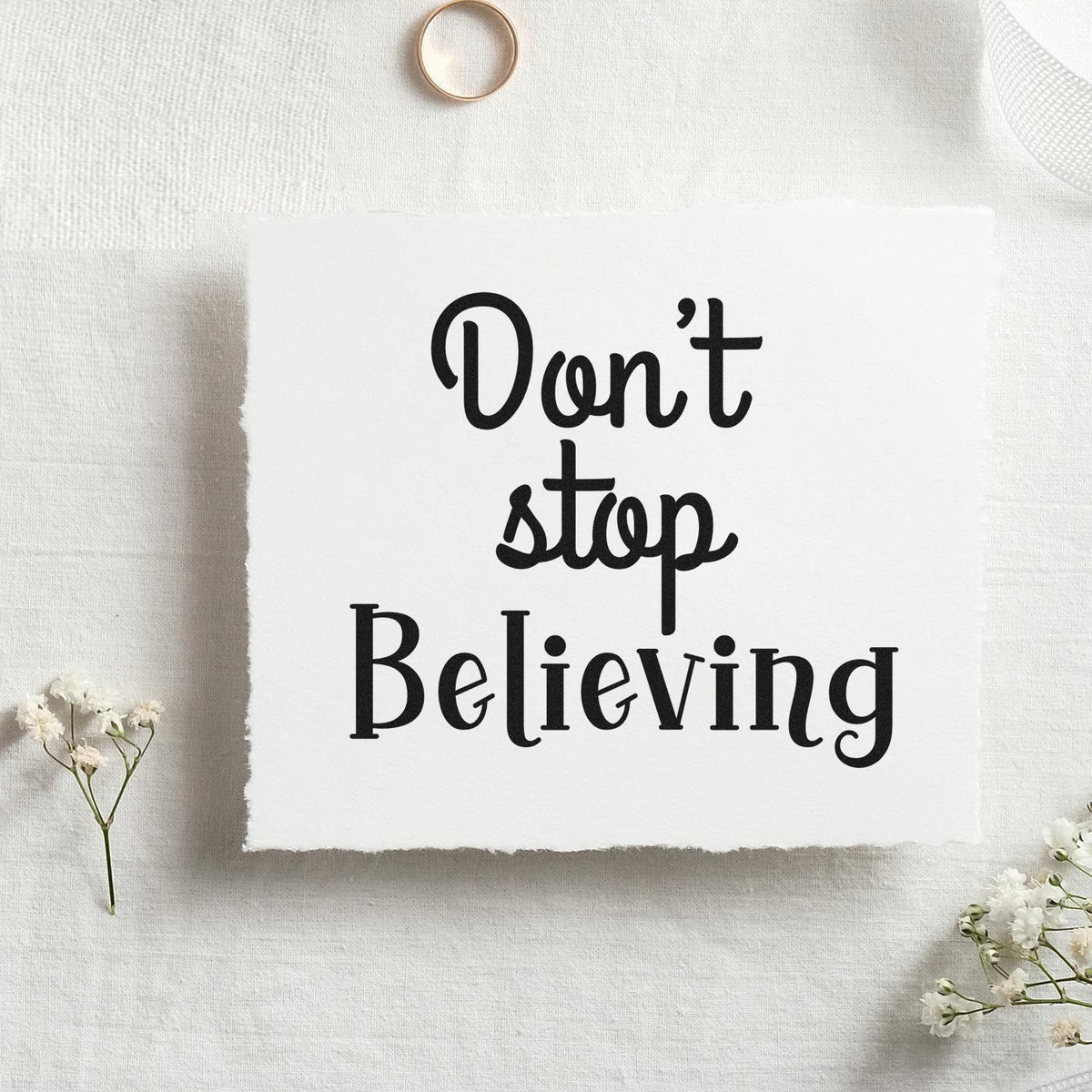 Black custom rubber stamp design reading Dont stop Believing on white textured paper with delicate white flowers.